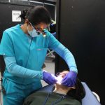 A dental specialist, wearing a mask and a light on her glasses, examines the inside of a patient's mouth.