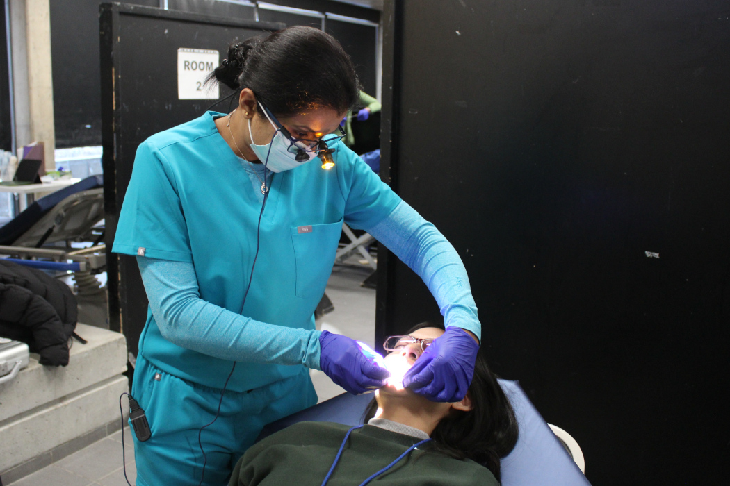 A dental specialist, wearing a mask and a light on her glasses, examines the inside of a patient's mouth.