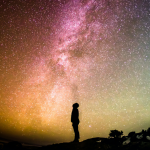 Person standing in the foreground just as a silouette and looking up to the stars. The sky and background are lit showing the milky ways in greens, reds, yellos and the clours inbetween. Thousands of stars across the night sky.