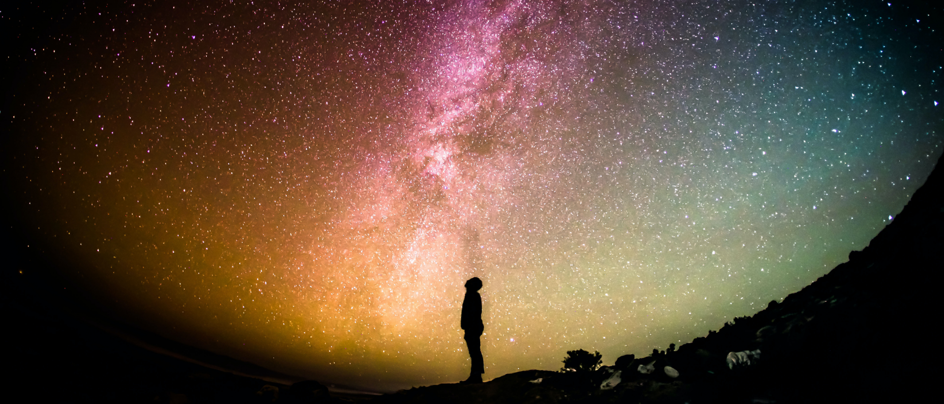 Person standing in the foreground just as a silouette and looking up to the stars. The sky and background are lit showing the milky ways in greens, reds, yellos and the clours inbetween. Thousands of stars across the night sky.