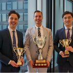 Three smiling law students wearing formal dress suits each hold a cup shaped trophy with handles.