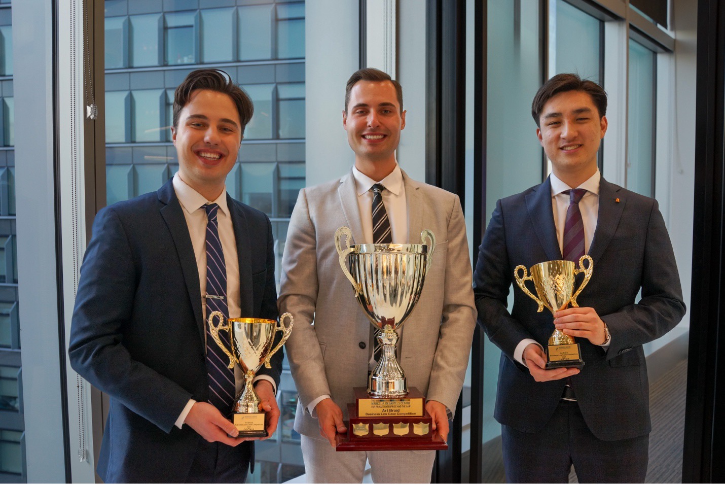 Three smiling law students wearing formal dress suits each hold a cup shaped trophy with handles.