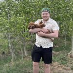 Man standing in front of brush of trees, wearing a backwards ballcap and holding a small brown dog.