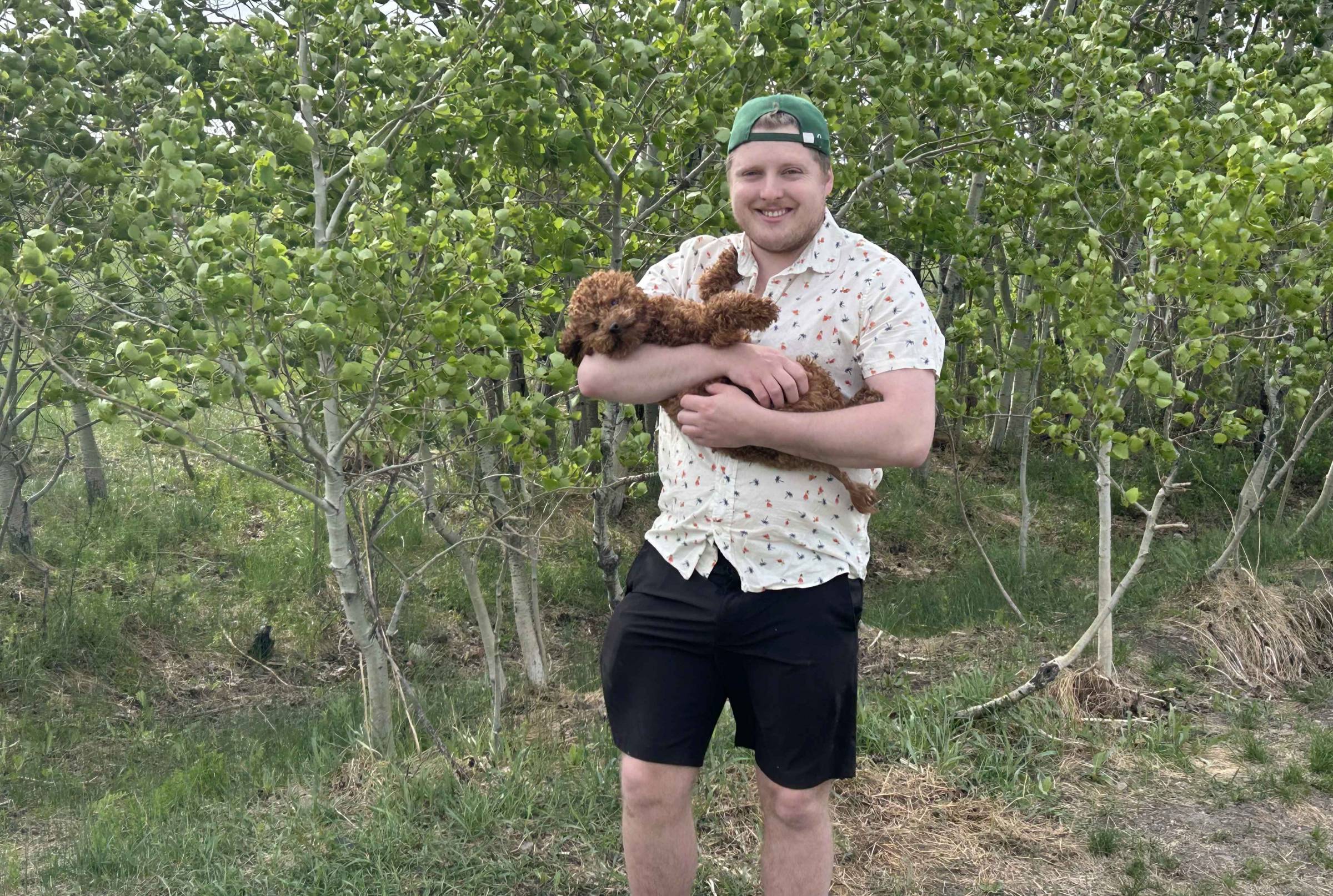 Man standing in front of brush of trees, wearing a backwards ballcap and holding a small brown dog.