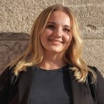 Headshot of Emma Winram wearing a black shirt and blazer, standing in front of a limestone wall, smiling at the camera.