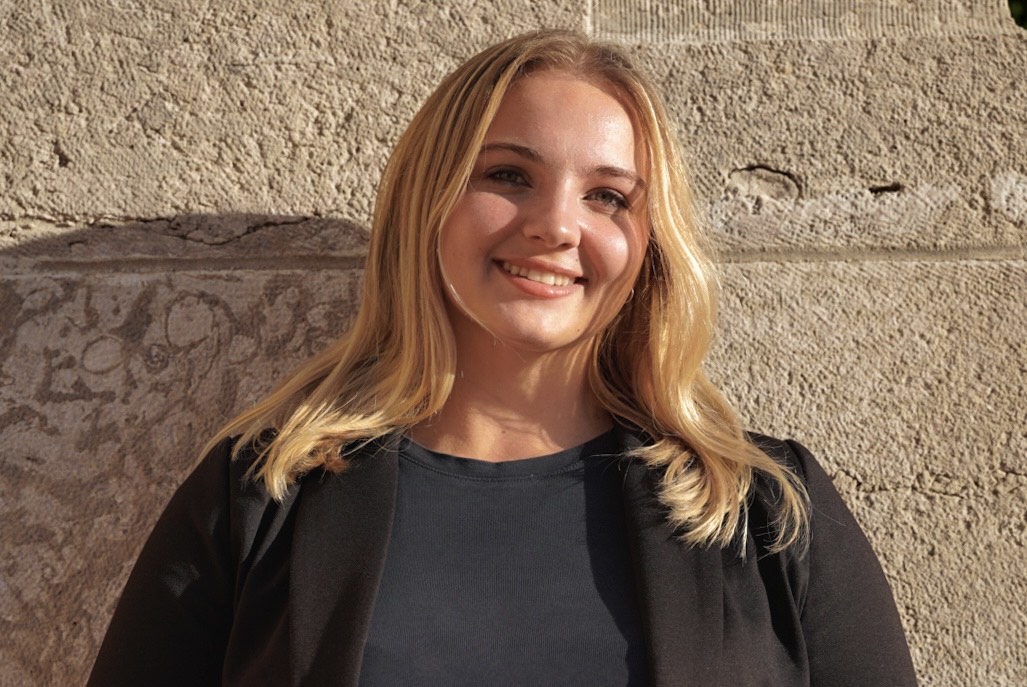 Headshot of Emma Winram wearing a black shirt and blazer, standing in front of a limestone wall, smiling at the camera.
