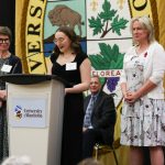 person at a white podium speaking into a mic with two people flanking them on their left and right.