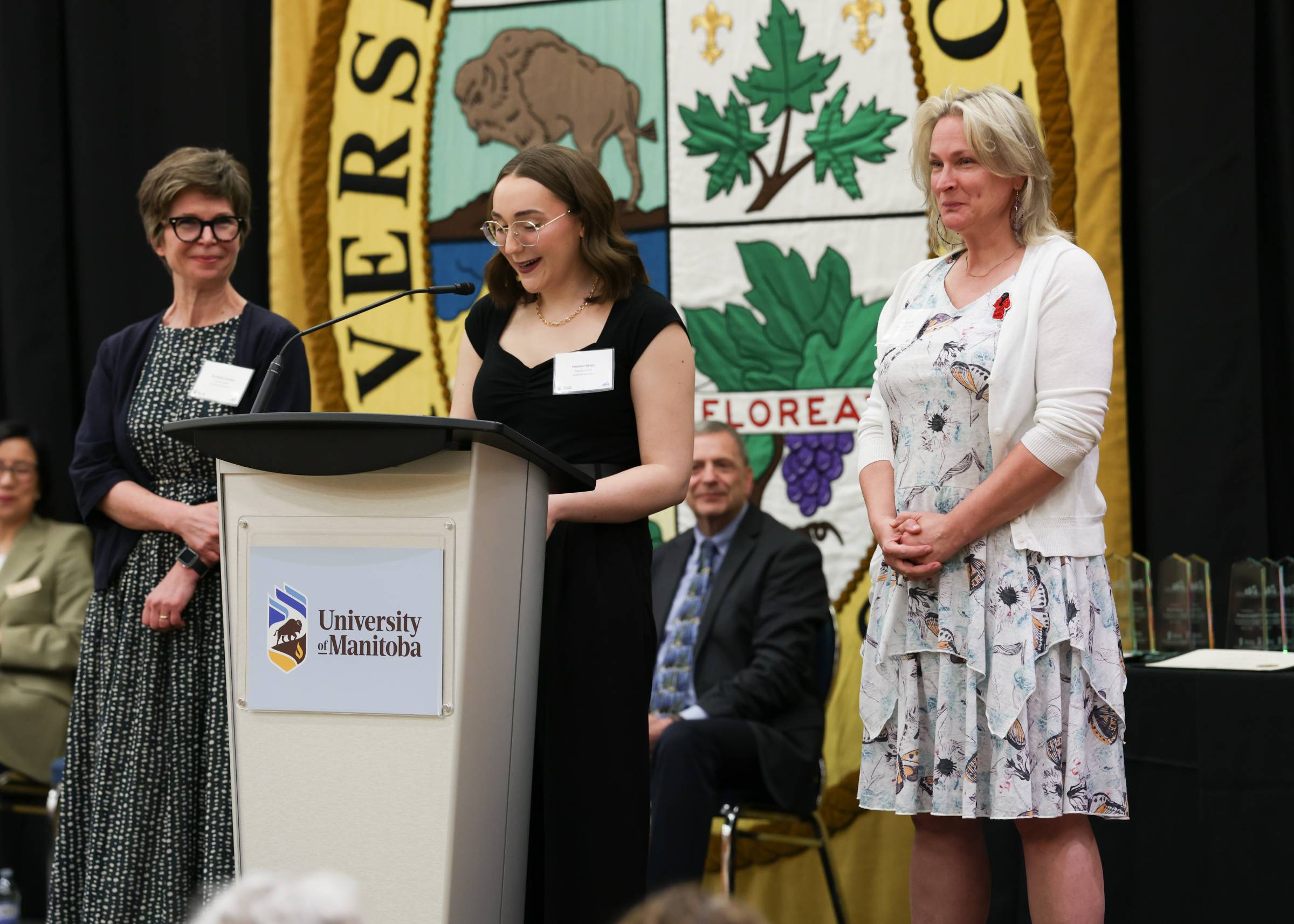 person at a white podium speaking into a mic with two people flanking them on their left and right.