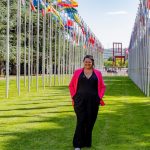 A woman standing in a grassy area with two rows of large white flagpoles with flags of various countries in a line on either side of her.