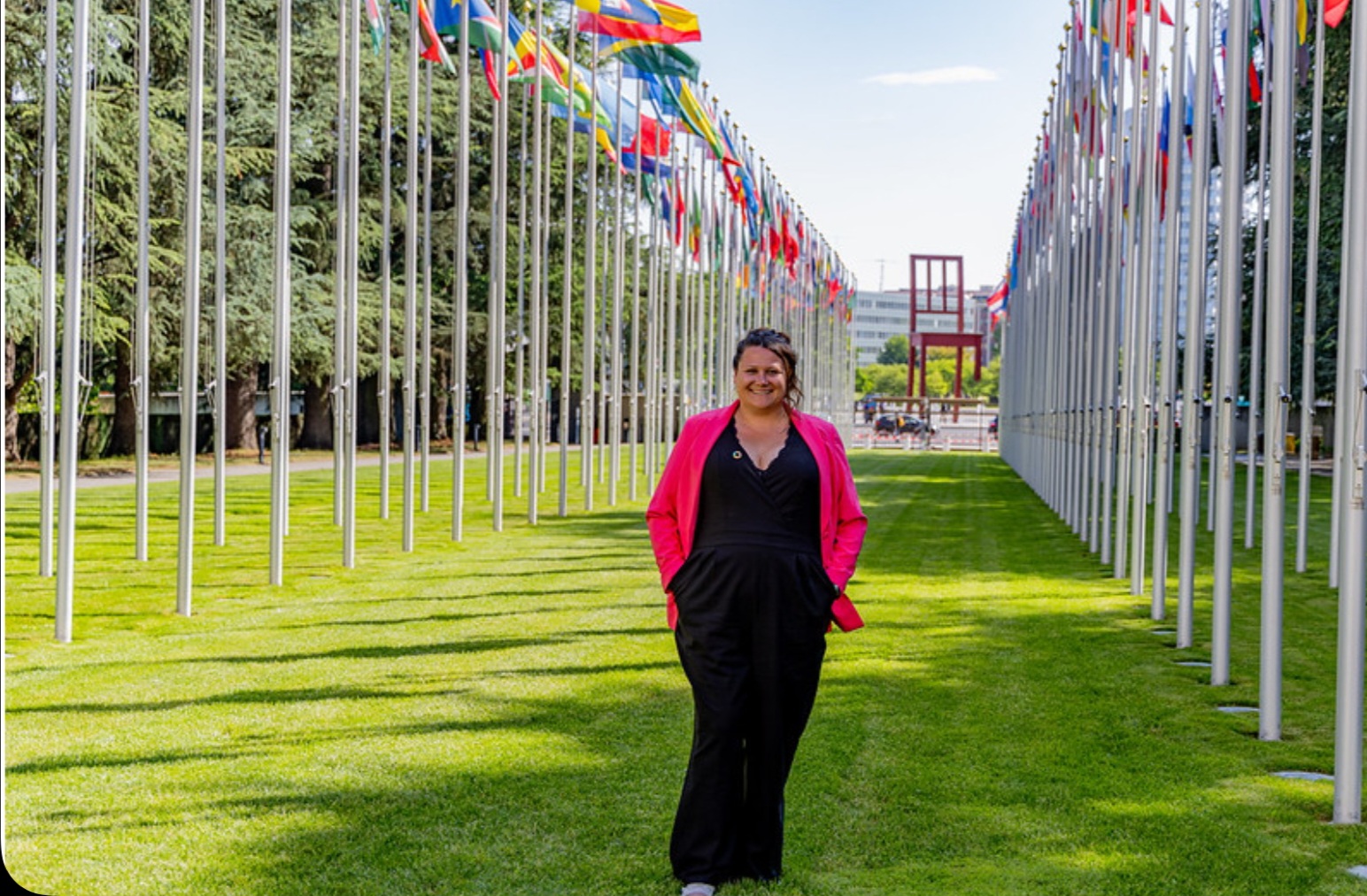 A woman standing in a grassy area with two rows of large white flagpoles with flags of various countries in a line on either side of her.
