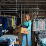 Kristie Pearson holding a box of clothes in a warehouse while looking at the camera