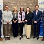 Seven law students in formal clothing stand smiling in front of two banners advertising the L. Kerry Vickar Business Law Clinic and the Manitoba Legal Clinic for the Arts.