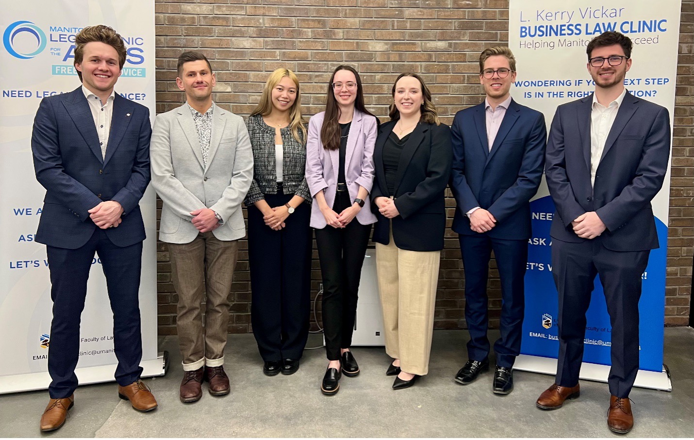 Seven law students in formal clothing stand smiling in front of two banners advertising the L. Kerry Vickar Business Law Clinic and the Manitoba Legal Clinic for the Arts.