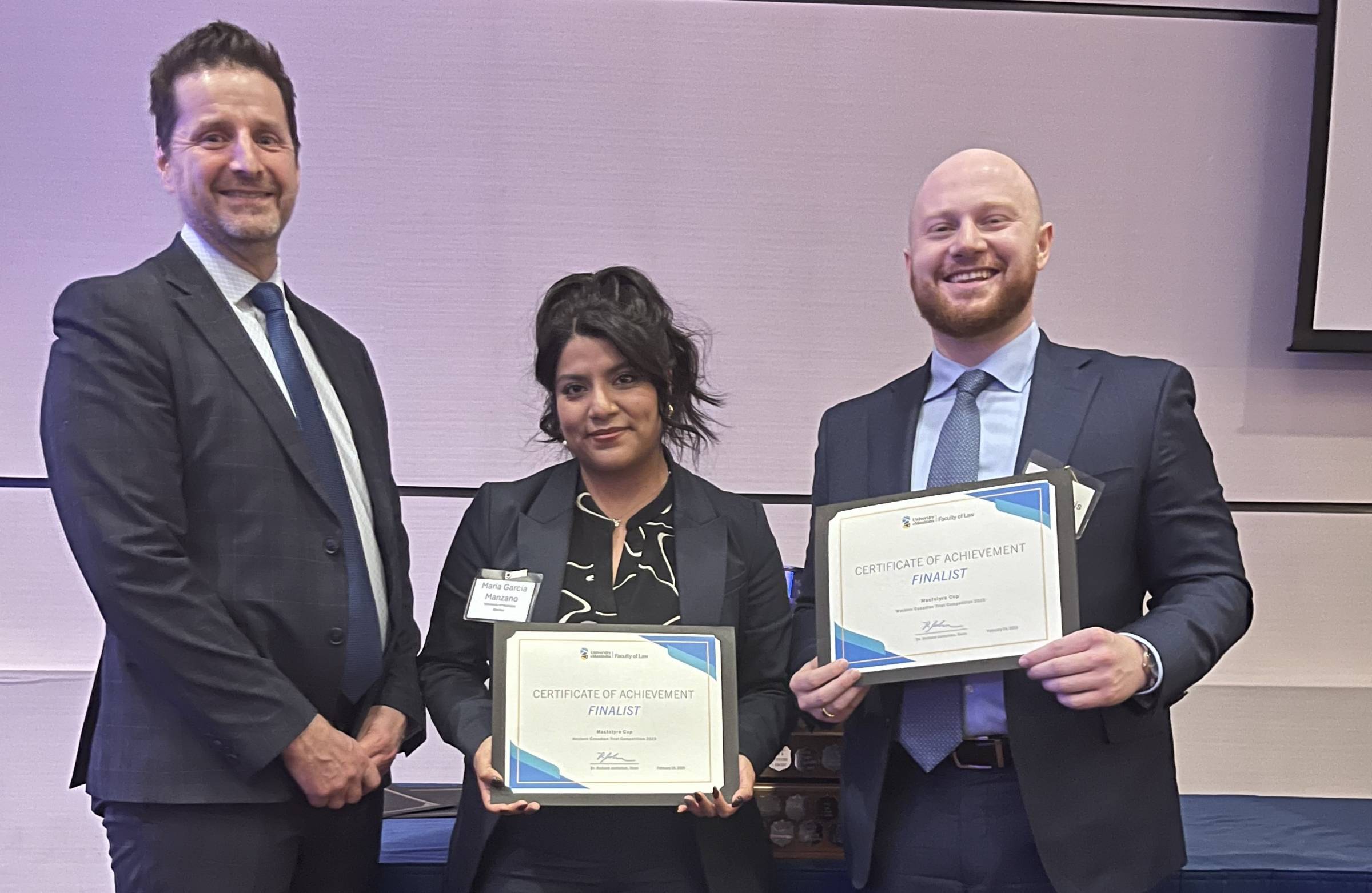 Three people smiling with two holding certificates indicating that they are finalists in the MacIntyre Moot Competition. Far left is Chief Judge of the Manitoba Provincial Court Ryan Rolston, then 3rd year law students Maria Garcia Manzano and Harlan Morris.