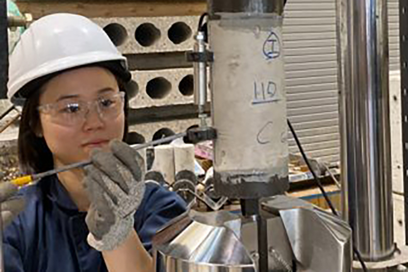 A person wearing a white hard hat, safety goggles, and gloves uses a tool to work on a cylindrical concrete sample in a lab setting. The setup includes industrial equipment and testing apparatus, with concrete blocks and testing machinery in the background."