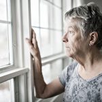 An elderly woman, peers out a window with a sad look on her face.
