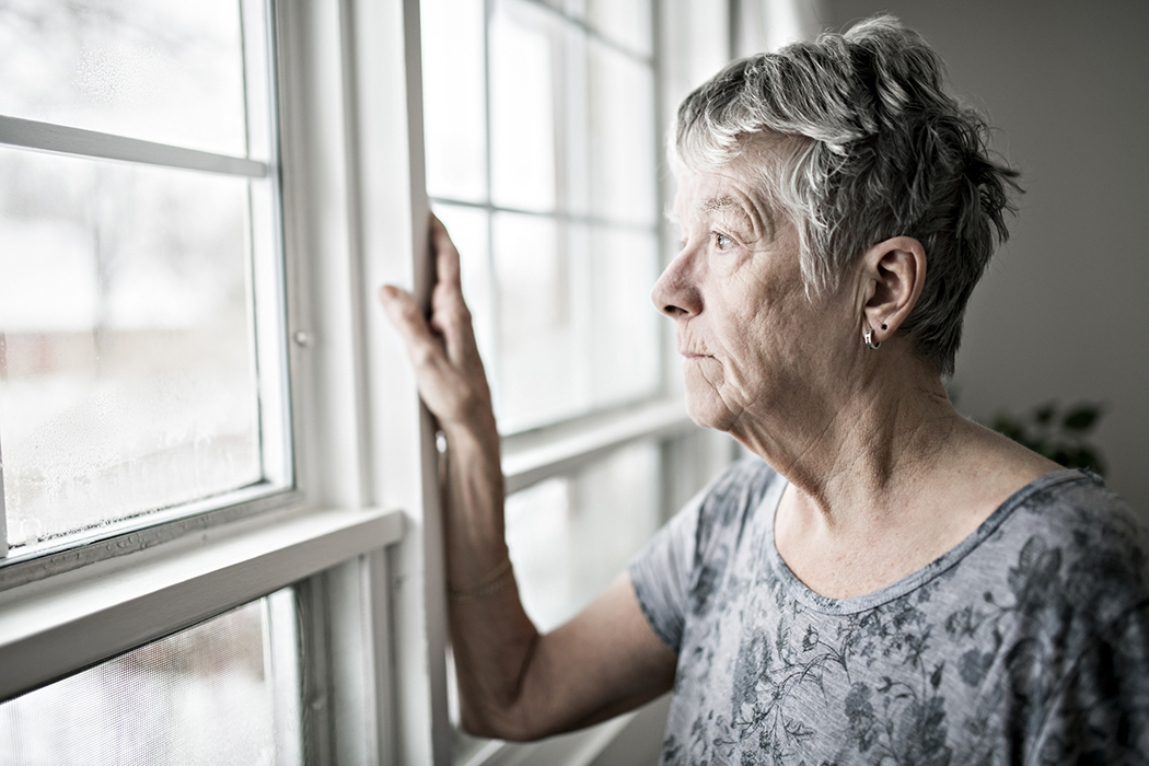 An elderly woman, peers out a window with a sad look on her face.