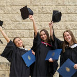 Three UM grads pose in front of a brick wall. They are holding their caps in the air in celebration.