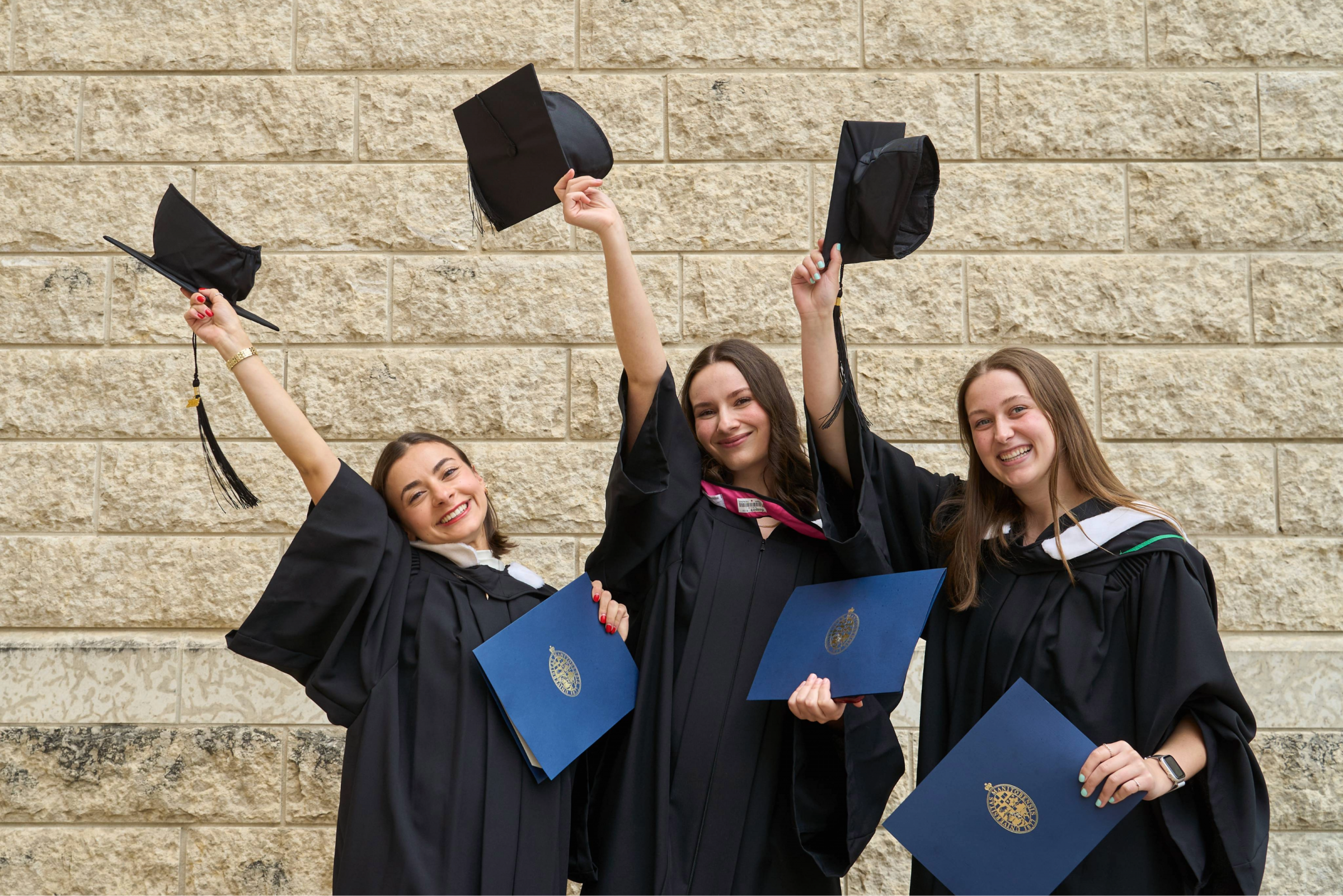 Three UM grads pose in front of a brick wall. They are holding their caps in the air in celebration.