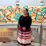 Kaydence Morgado Thomas smiles in front of mural with ribbon skirt