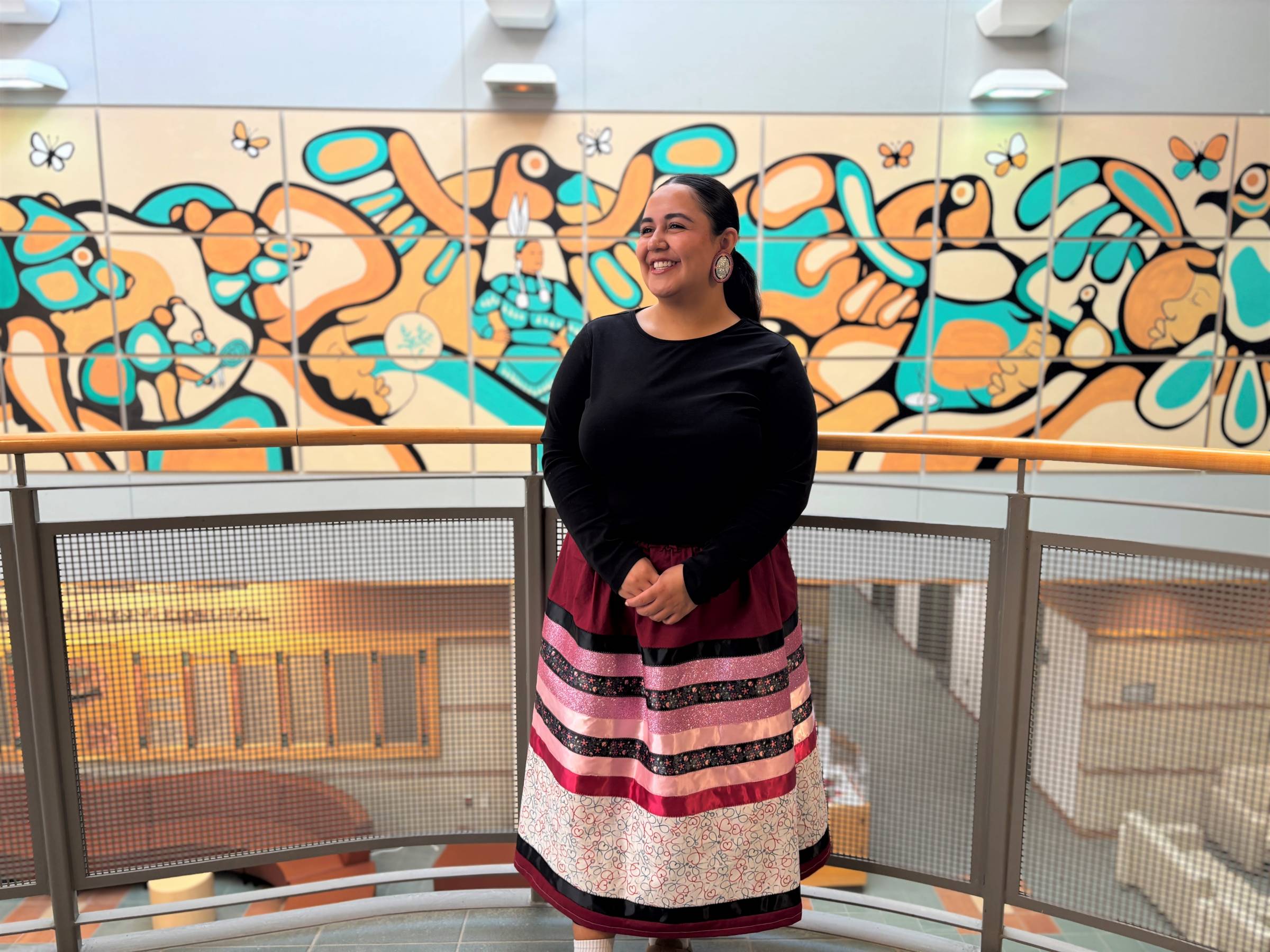 Kaydence Morgado Thomas smiles in front of mural with ribbon skirt