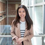 Riya Timpog standing against the metal and glass rail guards of the stairs in third floor of the engineering building with exterior brick walls in the background