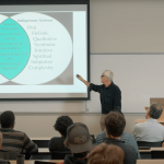 An instructor pointing at screen, talking to classroom of students