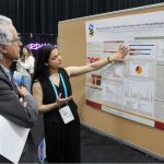 Navnoor places her hand on her poster, which is pinned to a bulletin board. A judge is listening to her and looking at the poster.