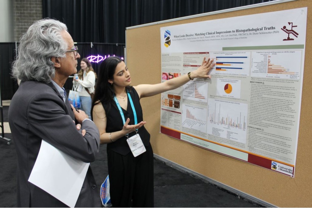 Navnoor places her hand on her poster, which is pinned to a bulletin board. A judge is listening to her and looking at the poster.