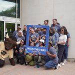 A group of people pose with Billy the Bison and a sign board reading "walk your way"