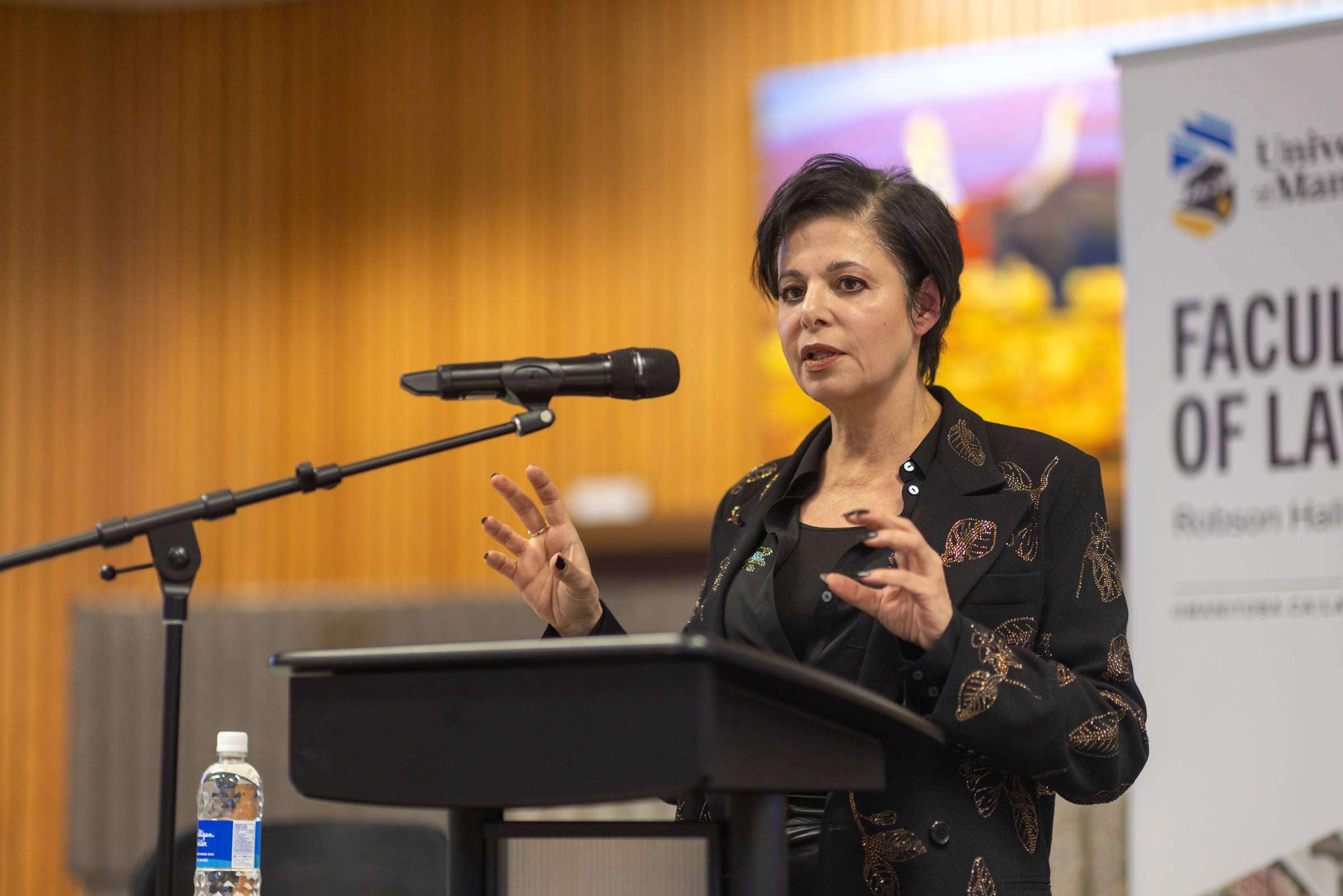 lawyer Marie Henein stands at a podium speaking into a mic with the Faculty of Law banner and moot courtroom art behind her. She is speaking and gesturing with her hands. Photo by Adam Dolman.