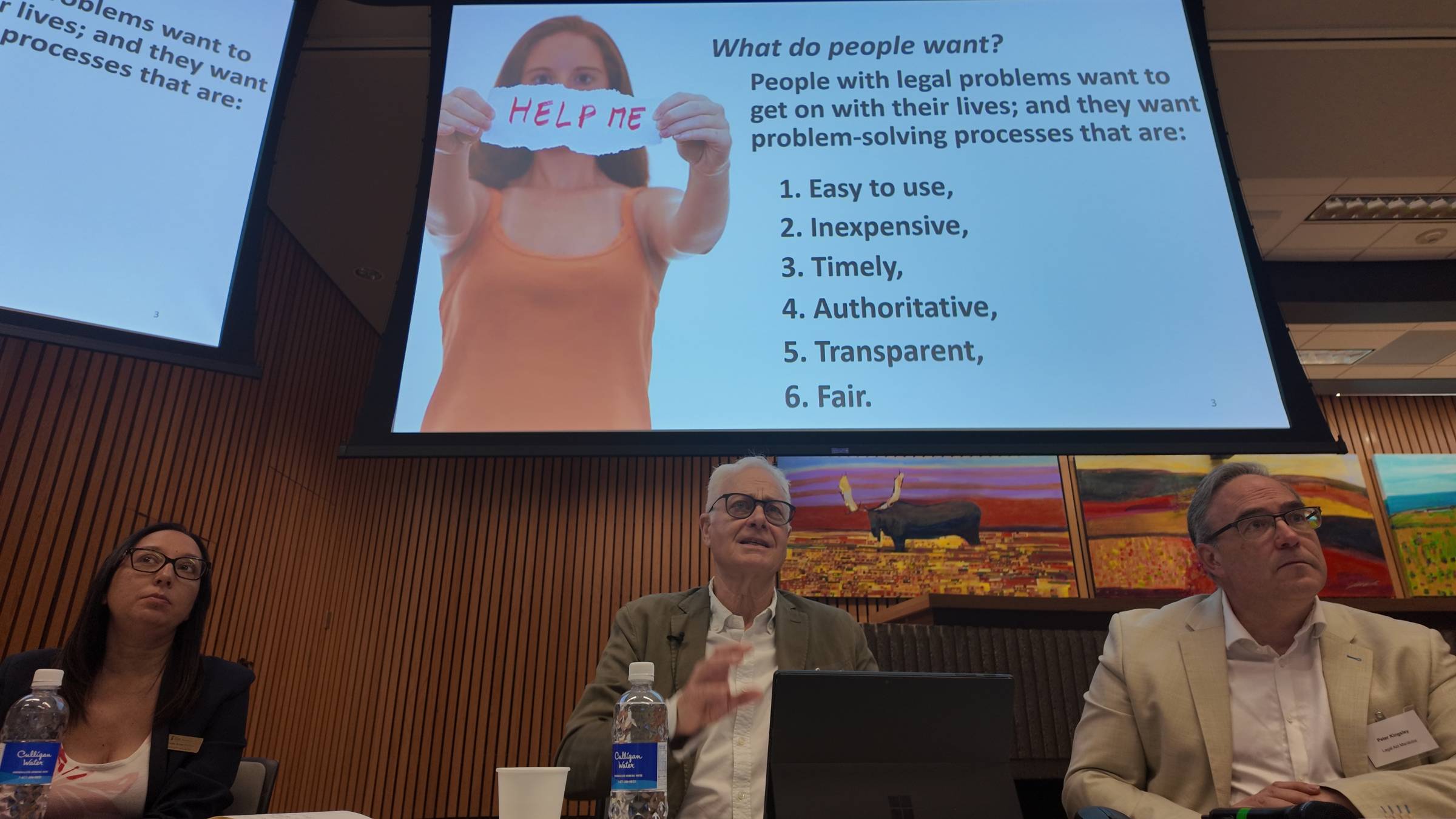 Three people sit under a screen showing a slide asking What do people want? People with legal problems want to get on with their lives; and they want problem-solving processes that are 1 easy to use, 2 Inexpensive 3 timely 4 authoritative 5 transparent 6 fair.