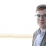 Headshot of Brandon Trask, a red-haired young man in glasses, blue checked shirt and grey suit jacket.