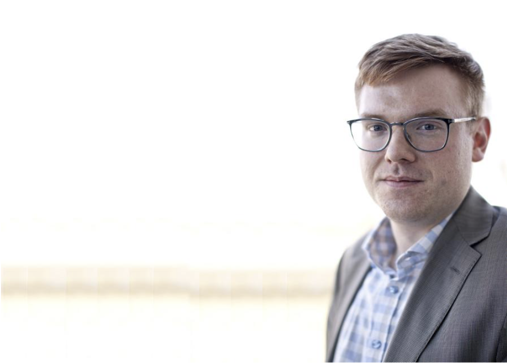 Headshot of Brandon Trask, a red-haired young man in glasses, blue checked shirt and grey suit jacket.