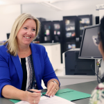 Two people seated at a desk in an office setting. Britt Kural, wearing a blue blazer, is holding a pen and reviewing travel health documents with the other person, who is partially visible and facing away from the camera.