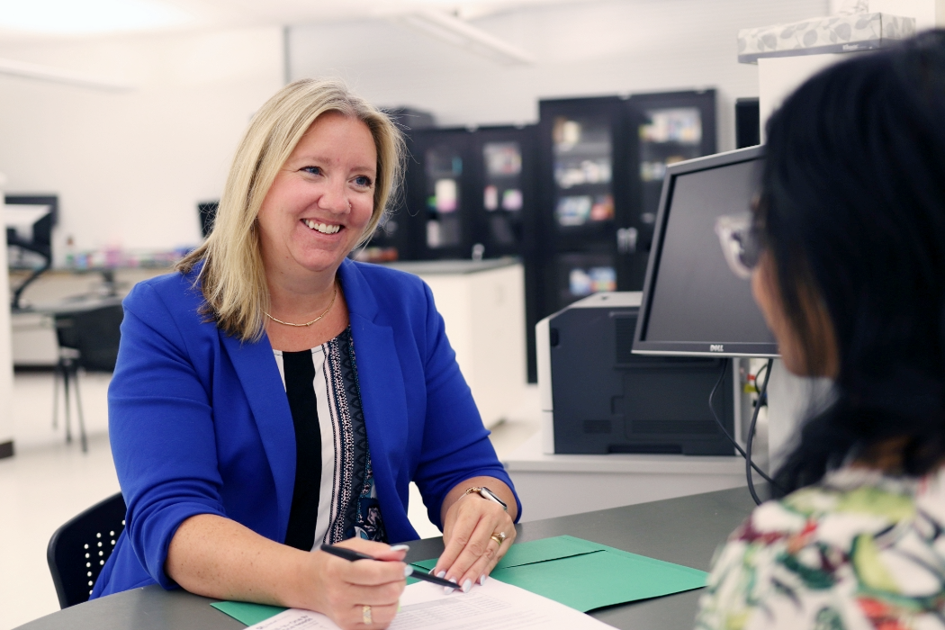 Two people seated at a desk in an office setting. Britt Kural, wearing a blue blazer, is holding a pen and reviewing travel health documents with the other person, who is partially visible and facing away from the camera.