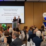 Two people stand behind a podium on a small stage. One speaks into a microphone. On a screen behind them text reads: "Welcome to the Department of Clinical Health Psychology Residency Program 55th Anniversary Celebration. Max Rady College of Medicine. Rady Faculty of Health Sciences." Dozens of people sit at round tables in the crowd.