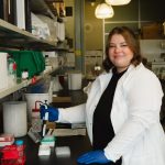 UM researcher, Courtney Marshall in a lab wearing a white lab coat and holding a lab apparatus while smiling at the camera.