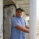 Man wearing a blue shirt and grey ball cap standing near the railing on a boat in Lake Winnipeg.