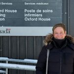 Dr. Judy Zetaruk stands in front of a building wearing a parka. A sign behind her reads: "Medical Services. Oxford House Nursing Station. Services medicaux. Poste de soins infirmiers Oxford House. Canada."