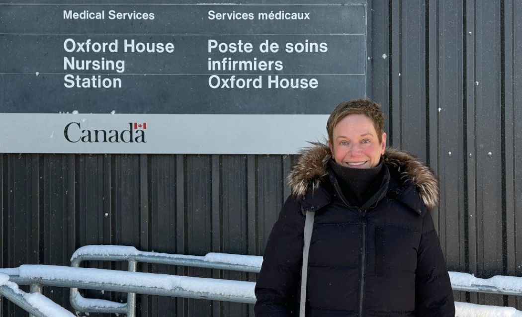 Dr. Judy Zetaruk stands in front of a building wearing a parka. A sign behind her reads: "Medical Services. Oxford House Nursing Station. Services medicaux. Poste de soins infirmiers Oxford House. Canada."