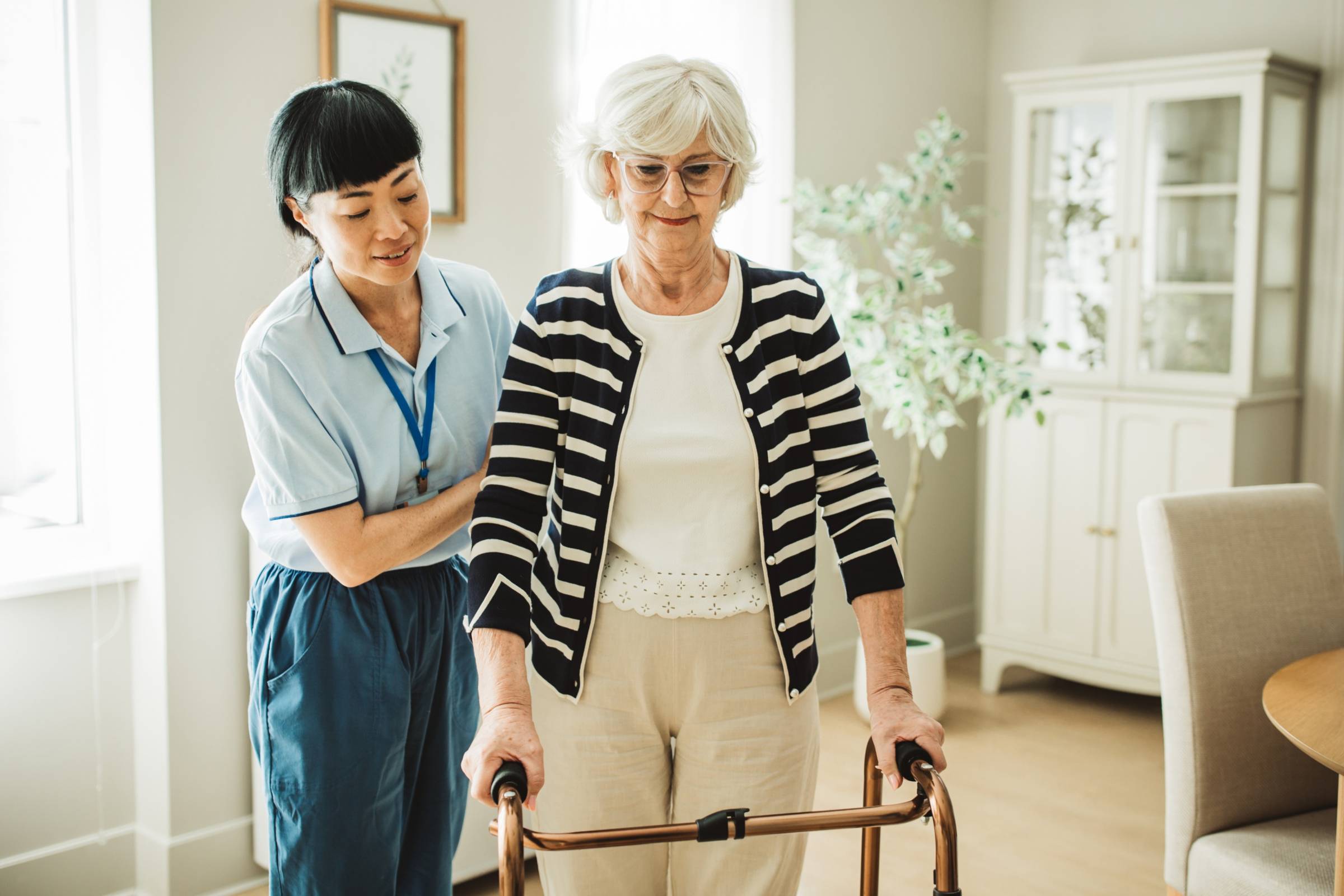 A person helps an older person while they walk using a walker in a house.