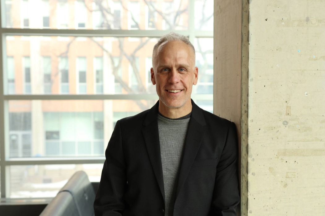 Dr. Jamie Falk smiles at the camera while leaning on a pillar inside the Apotex Centre, with the building’s exterior visible through the windows in the background.
