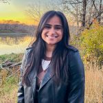 A woman standing on a river bank in the fall, wearing a black, leather jacket and smiling at the camera.