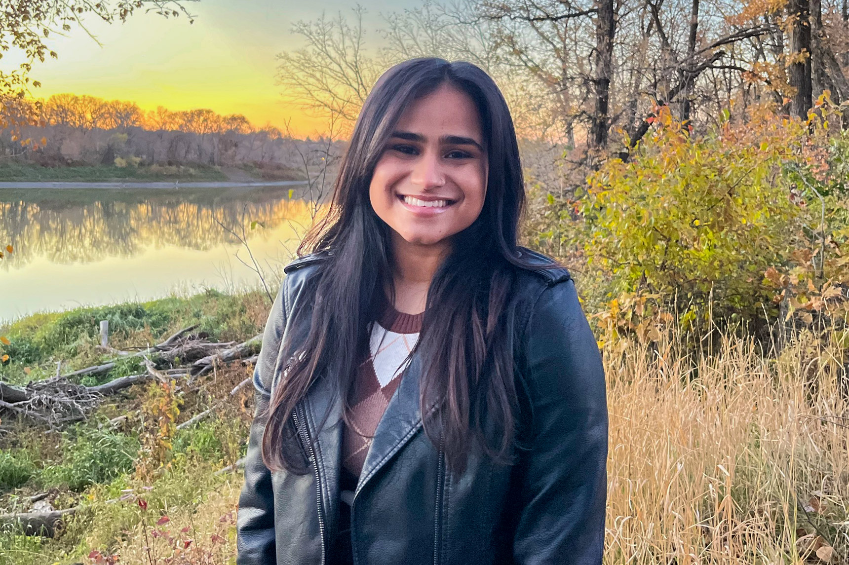A woman standing on a river bank in the fall, wearing a black, leather jacket and smiling at the camera.
