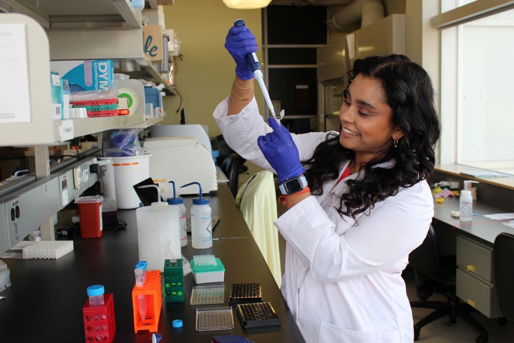 Tamarah Singh works with a pipette in a lab. She is wearing gloves and a lab coat.