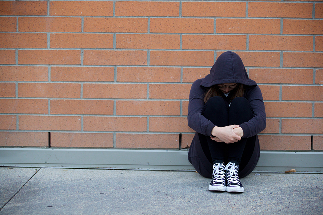 A young person sits on a sidewalk against a wall, head down.