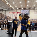 University of Manitoba president and the Bisons mascot stand on stage in front of a packed gymnasium of cheering students during a pep rally. Both wear ‘We Are All Bisons’ shirts, and the crowd holds gold rally towels. A large screen in the background displays the audience.