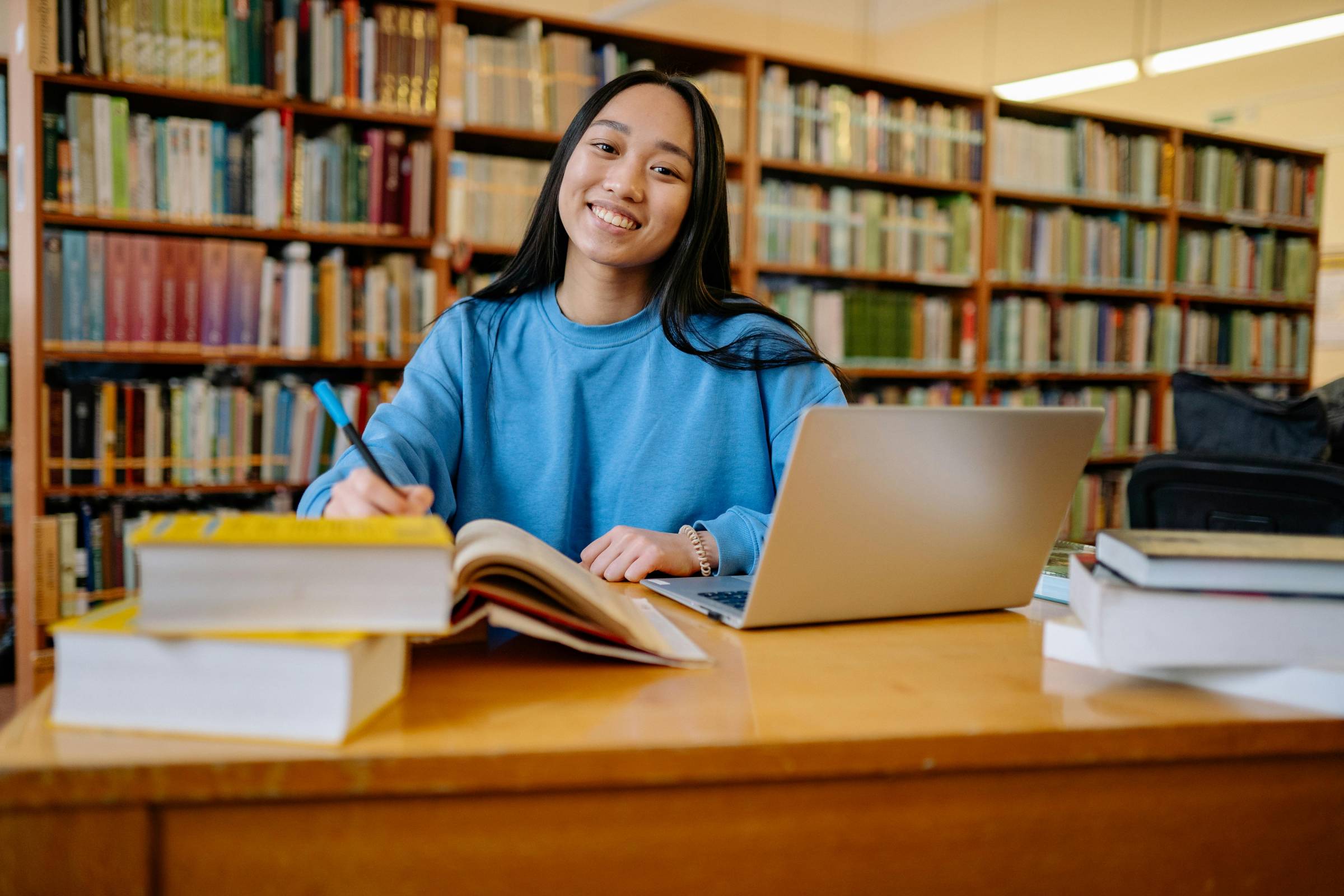 Student in a library with a laptop open and a pile of books open to the left.