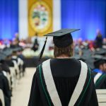 A grad in cap and gown walks down the centre aisle at Convocation. A banner with the University Crest hangs on the wall in the distance.